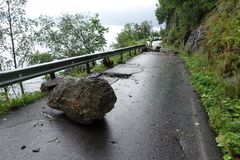 En smal asfaltert vei med autovern. En stor stein ligger midt i veien. På ene siden er det en bratt fjellside, på andre siden av autovernet er trær i en skråning. En person i gul jakke står lenger bak i bildet sammen med biler i kø.