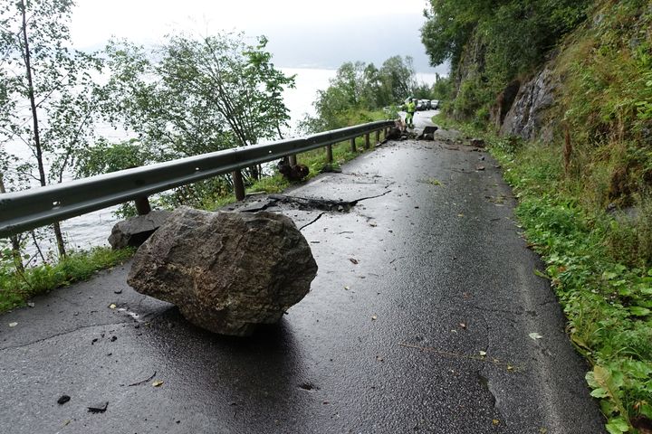 En smal asfaltert vei med autovern. En stor stein ligger midt i veien. På ene siden er det en bratt fjellside, på andre siden av autovernet er trær i en skråning. En person i gul jakke står lenger bak i bildet sammen med biler i kø.