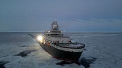 FF Kronprins Haakon bryter gjennom tynn havis under et blågrått himmellys. Skipet er opplyst av sterke arbeidlys, og et langt spor i isen viser ruten det har presset seg gjennom. Isflak og mørke råker omgir forskningsfartøyet i det arktiske landskapet.