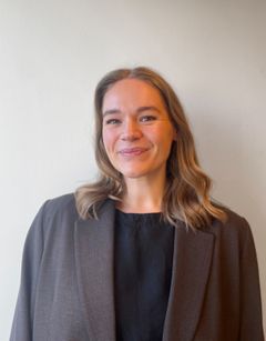 Person i brun jakke og svart topp smiler mot kameraet.