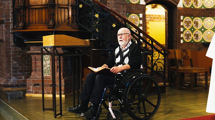 Tekstlesing i kirke, tekstleseren er en mann i rullestol. Gulldekor på veggen bak, lesepult ved siden av mannen.