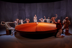 Liggende bilde fra danseforestillingen KØTER av Dansk Danseteater. 10 dansere står rundt en stor vifteform. En danser sitt hodet stikkende opp i midten av viften.