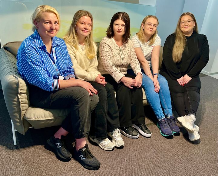 Fem, blide, norske kundesentermedarbeidere som sitter ved siden av hverandre i en sofa.