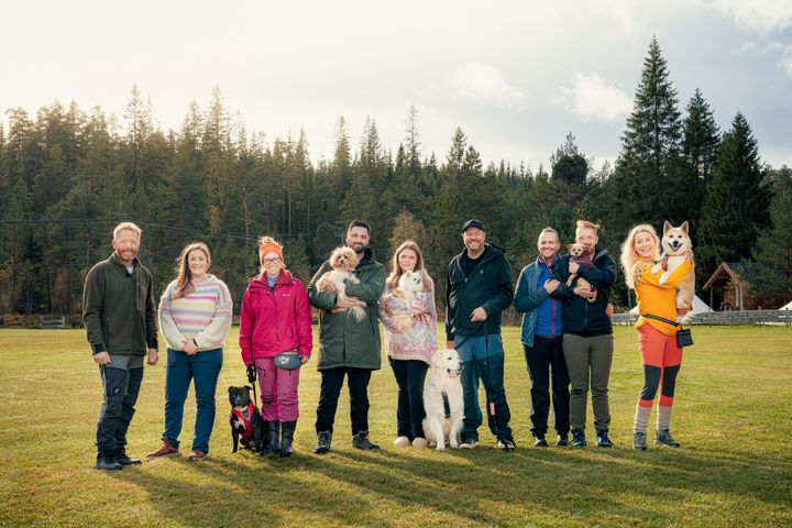Gruppebilde av ni mennesker med seks hunder på en åpen gressplen foran en skog.