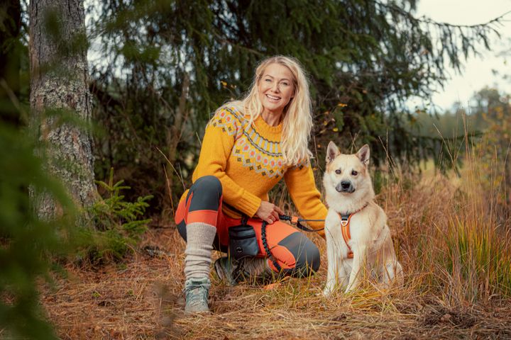 En%20kvinne%20kledd%20i%20fargerike%20kl%E6r%20sitter%20i%20skogen%20med%20en%20hvit%20hund%20ved%20sin%20side.