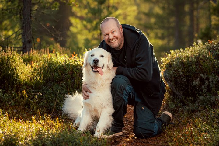 En%20smilende%20mann%20som%20sitter%20p%E5%20huk%20i%20skogen%20ved%20siden%20av%20en%20glad%2C%20hvit%20hund.