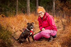 Bilde av en kvinne som sitter på huk ved siden av en hund i skogen. Hun har på seg en rød jakke og rosa bukser.