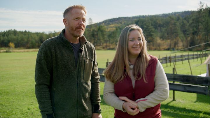 En%20mann%20og%20en%20kvinne%20st%E5r%20utend%F8rs%20p%E5%20en%20gressplen%20med%20tr%E6r%20og%20en%20bl%E5%20himmel%20i%20bakgrunnen.