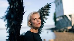 Kvinne med svarte vinger på en strand ser inn i kameraet