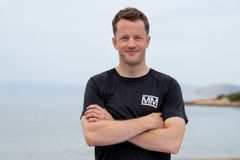 Tarjei Bø i svart Mesternes mester-drakt står med armene i kors ved havet, smilende mot kameraet.