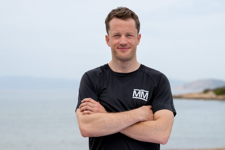 Tarjei%20B%F8%20i%20svart%20Mesternes%20mester-drakt%20st%E5r%20med%20armene%20i%20kors%20ved%20havet%2C%20smilende%20mot%20kameraet.