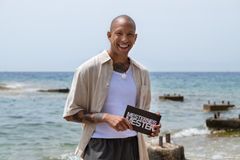 Tete Lidbom smiler og holder et «Mesternes mester» manuskort, stående med havet i bakgrunnen