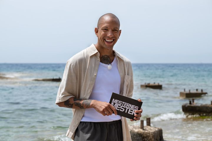 Tete%20Lidbom%20smiler%20og%20holder%20et%20%ABMesternes%20mester%BB%20manuskort%2C%20st%E5ende%20med%20havet%20i%20bakgrunnen