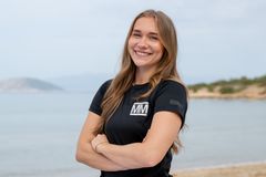 Julie Thorsen smiler med armene i kors på en strand iført svart Mesternes mester-drakt, med hav og fjell i bakgrunnen.