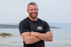 MMA-utøver Emil Meek smiler med armene i kors, iført svart Mesternes mester-drakt,  med hav og kystlinje i bakgrunnen.