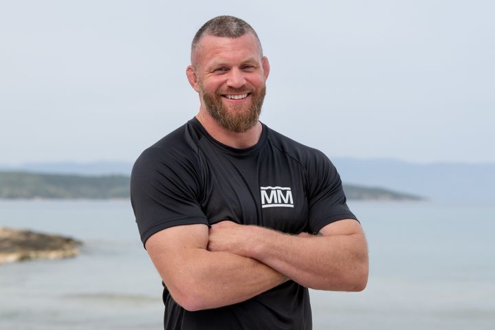 MMA-ut%F8ver%20Emil%20Meek%20smiler%20med%20armene%20i%20kors%2C%20if%F8rt%20svart%20Mesternes%20mester-drakt%2C%20%20med%20hav%20og%20kystlinje%20i%20bakgrunnen.