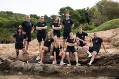 Gruppebilde av de ti deltakerne i «Mesternes mester» 2027, iført svarte Mesternes mester-drakter, samlet på klipper ved en strand i Porto Heli, Hellas.