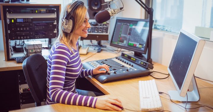 Kvinnelig radioprogramleder sitter ved pult med miksebord, mikrofon og pc