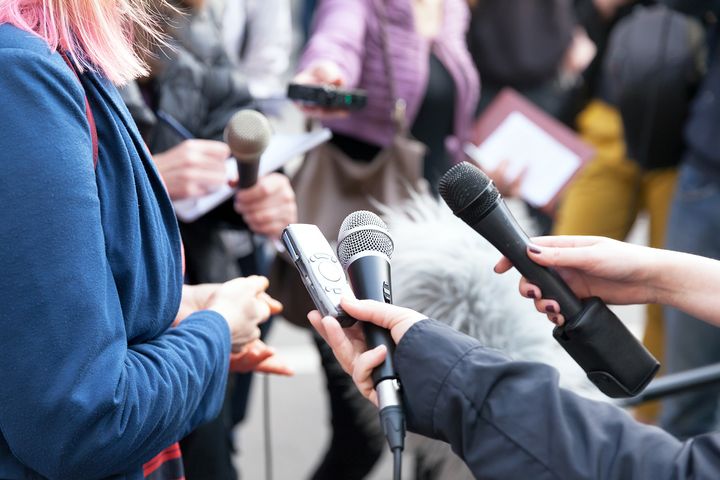 Nærbilde av hender som holder hver sin mikrofon. Vi ser ingen ansikter, og intervjuobjekt og journalister i halvsirkel rundt er uskarpe rundt mikrofonene.