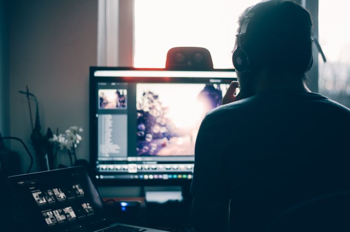 Mann sitter ved arbeidsplass og redigerer. Vi ser mannen bakfra. Han har på seg hodetelefoner. Han har en laptop til venstre for seg og en større skjerm foran.