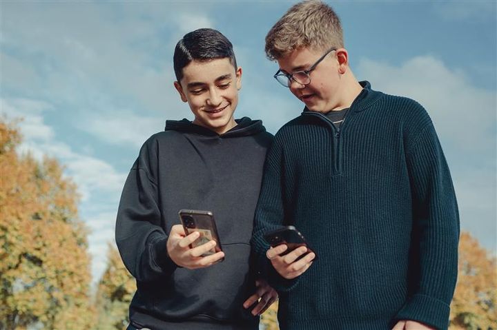 To gutter står skulder ved skulder og ser på mobilene sine og viser hverandre. I bakgrunnen er det trær og lettskyet himmel.