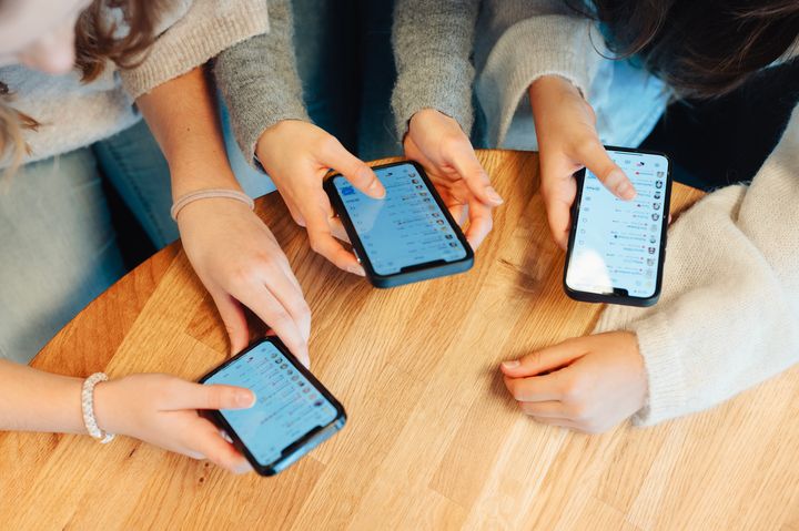 Nærbilde av hender og mobiltelefoner. Tre jenter sitter ved et bord. Vi ser at alle har Snapchat oppe på telefonskjermen.