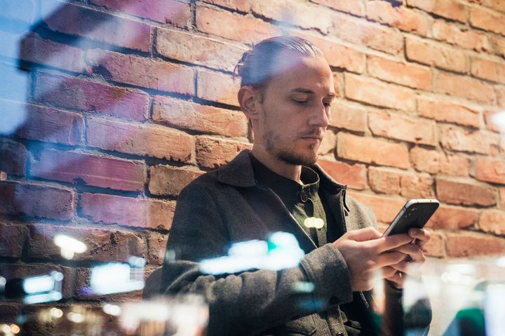 En ung mann står med ryggen mot en vegg av teglsten og ser på mobiltelefonen sin. Bildet er tatt gjennom et vindu, og det er refleksjoner av lys i glasset.