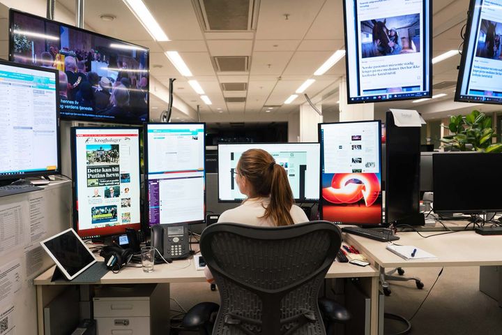 En kvinnelig journalist fotografert bakfra ved desken. Hun har mange skjermer foran seg.