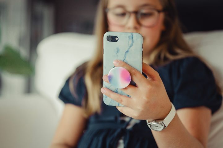 Barn sitter i sofaen og holder en smarttelefon med blåmarmorert deksel og fargerik popsocket. Barnet har briller og klokke, og ser på skjermen.