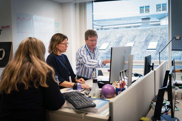 Tre personar står ved ein pult i eit redaksjonslokale og ser mot ein pc-skjerm. Den ene peiker mot skjermen.