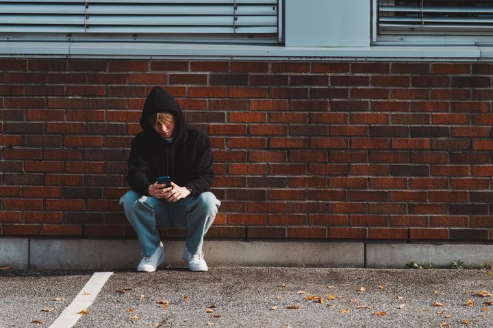 En tenåringsgutt med olabukse og hettegenser sitter på huk med ryggen mot en rød murstein-vegg. Han ser på mobiltelefonen sin.