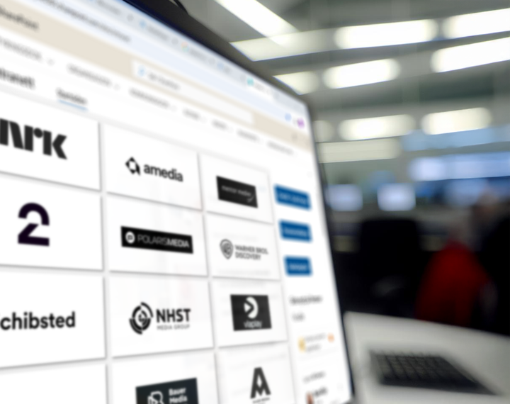 Dataskjerm med oversikt over norske mediehus, inkludert logoene til NRK, TV 2, Schibsted, Amedia, Polaris Media, NHST Media Group, Warner Bros. Discovery og flere. Bildet er tatt i et kontormiljø med uskarp bakgrunn.
