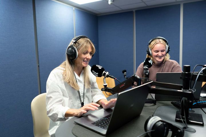 To kvinner sitter ved et bord i et studio for lydopptak. De har på hodetelefoner, og foran seg har de hver sin mikrofon og pc.