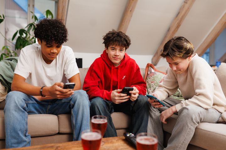 Tre tenåringsgutter sitter i en lys sofa og ser på hver sin telefon. På bordet foran dem står tre glass rød saft.