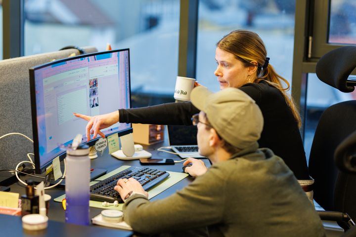 To personer sitter ved en pc. Mann nærmest har briller, grønn skjorte og caps. Kvinne lengst fra kamera har svart genser, hun holder en kopp i den ene hånden og peker på skjermen med den andre.