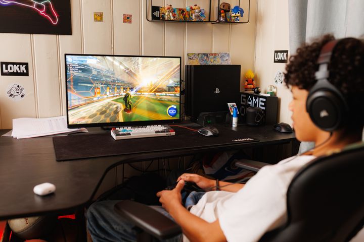 En gutt tidlig i tenårene sitter tilbakelent i gamingstol og spiller. Han er avbildet på skrå bakfra. Han har hvit t-skjorte og mørkt hår. I hendene har han en kontroller og skjermen er på en pult foran ham.