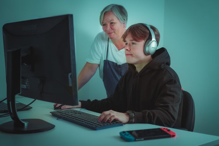 En tenåringsgutt sitter foran PC mens en voksen kvinne står ved siden av og følger med på skjermen. De er fotografert skrått forfra, slik at det ikke vises hva de ser på skjermen.