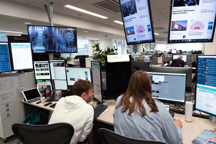 Frå avisredaksjon. Nærast kamera sit to personar med ryggen til kamera ved ein pult med mange skjermer. Lenger frå kamera er det mange pultar og pc-skjermar der det sit folk og jobbar.