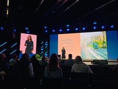 En kvinne presenterer på en scene med en stor skjerm i bakgrunnen. Publikum sitter i salen og ser på presentasjonen.