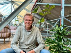 Bildet viser en smilende Ole André Sivertsen som sitter innendørs i et rom med store vinduer og mange grønne planter rundt.