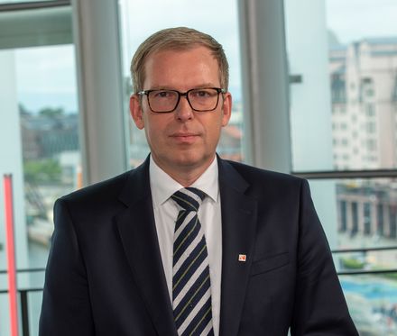 En mann i dress med briller står foran et stort vindu.