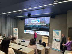 Kvinne presenterer på en teknologikonferanse med publikum og stor skjerm i bakgrunnen.