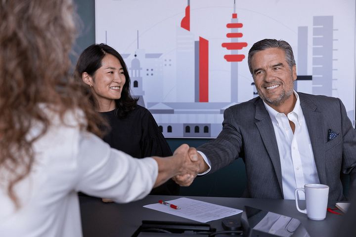 En person håndhilser med to smilende personer ved et bord med papirer.
