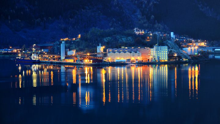 Nattbilde av Odda med opplyste bygninger langs fjorden og lysrefleksjoner i vannet, omgitt av mørke fjell.