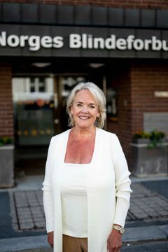 En smilende kvinne i hvit topp og jakke står foran døren til Norges Blindeforbund.