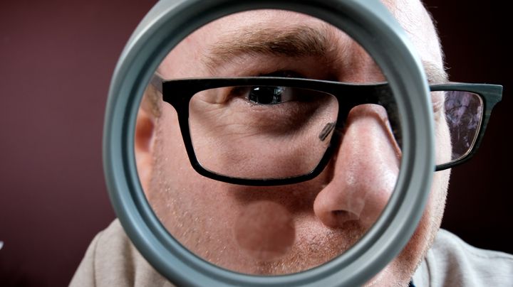 Nærbilde av mann som holder et forstørrelsesglass foran øyet. Han har briller på.