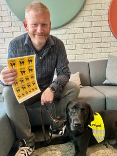 En smilende mann holder opp et ark med gule klistremerker som det sår førerhund velkommen på. Ved siden av han ligger en svart labrador som har refleksvest som det står førerhund på.