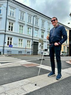 En mann med hvit stokk står oppå en linje med lyse, opphøyde heller som er lagt i asfalten på fortauet.