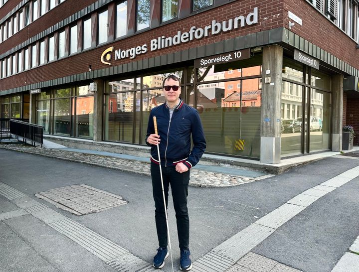 En mann med hvit stokk står på et fortau som det er lagt en linje med lyse heller i.