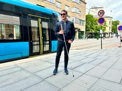 En mann med hvit stokk står på en trikkeholdeplass ved noen heller med opphøyde linjer på seg.